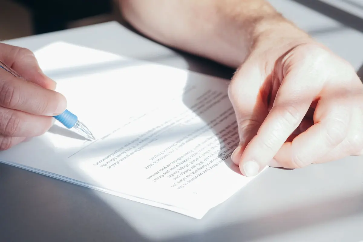 Two hands on a printed page, one holding a blue pen over the text